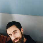 Close-up portrait of a bearded man with green eyes posing indoors against a blue wall.